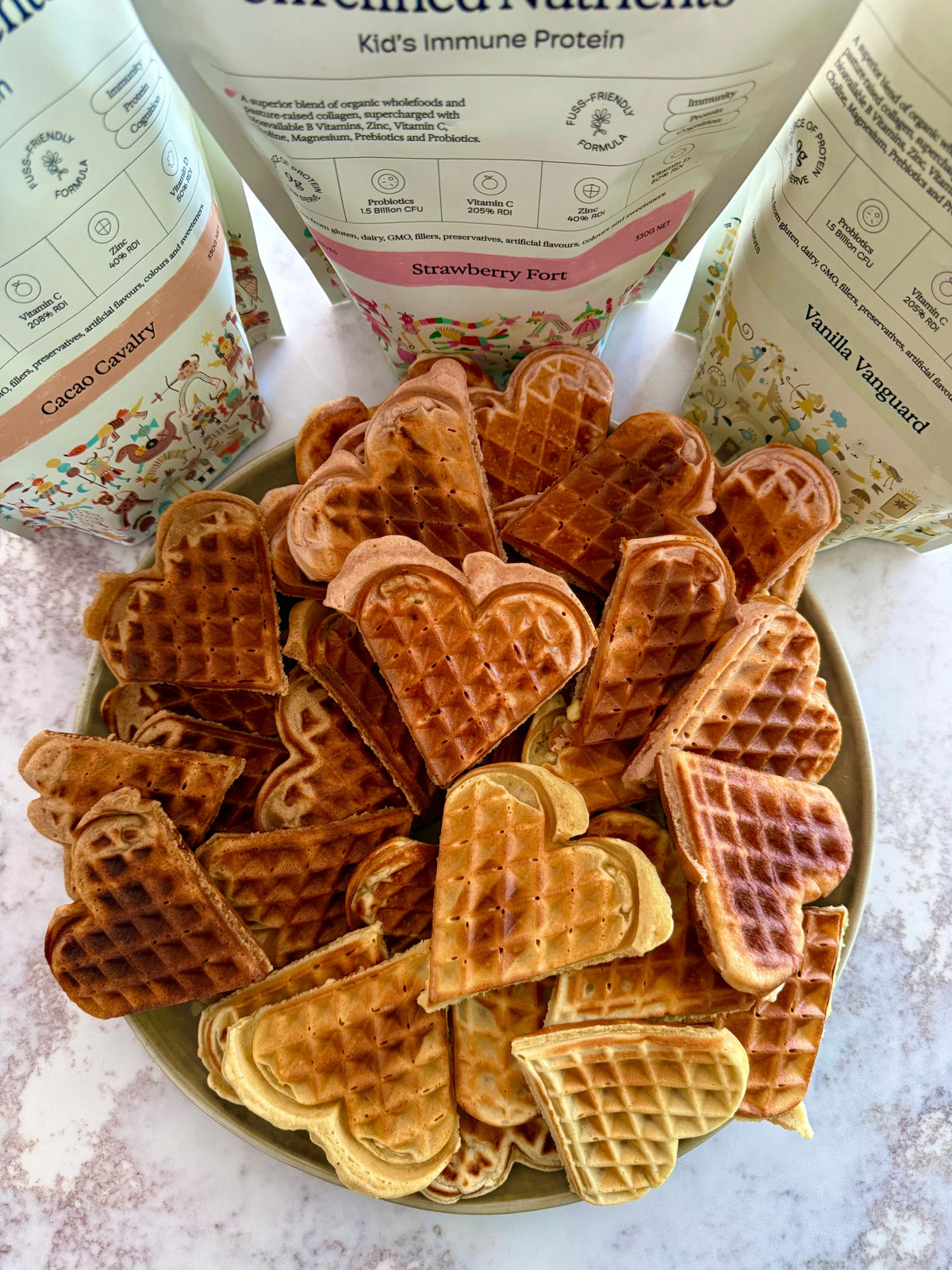 Plate of heart-shaped protein waffles in front of Kids Immune Protein in Cacao Cavalry, Strawberry Fort and Vanilla Vanguard